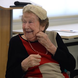 An elderly woman smiling as she wears a cream coloured pillbox hat. She is wearing a beaded necklace and a long sleeve black and red top.
