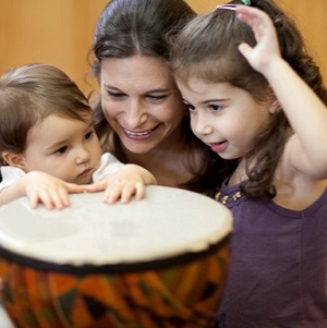 A woman smiles as two children either side of her play a drum