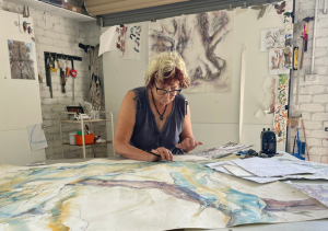 Artist sits at desk painting. The artwork shows blue and purple tones on a large piece of paper.