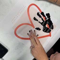 Baby's hand making a handprint on a piece of paper with a pink heart on it.