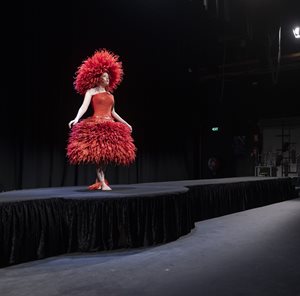 Model wearing a red feather hat and dress on a black room