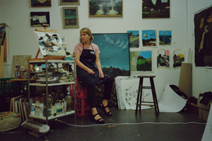 artist in her own studio, sitting down on a red stall surrounded by landscape paintings